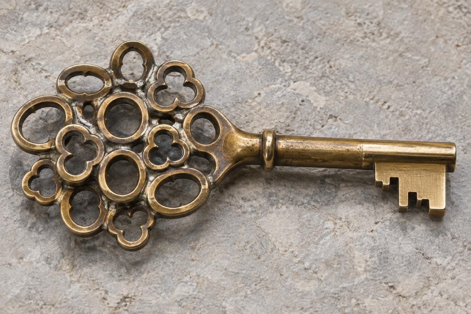 A brass key lies on pale stone. Twelve overlapping, worn key heads are fused into a broad cluster that narrows into one shaft and a single bright, toothed blade.