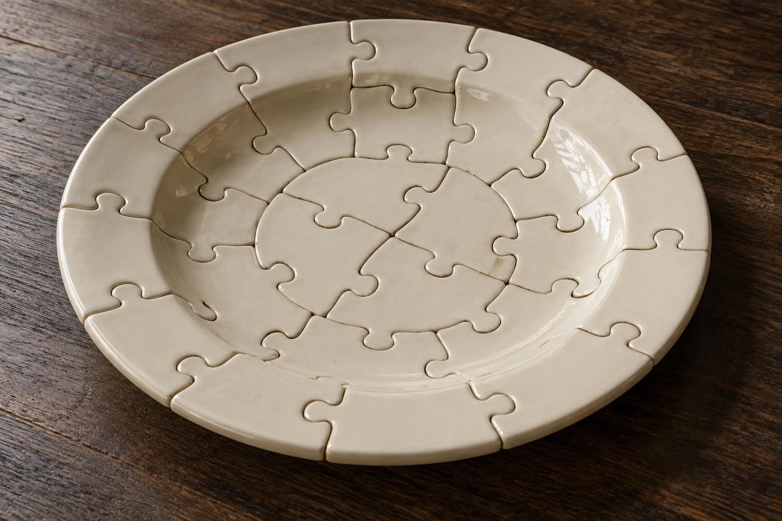 An ivory porcelain dinner plate assembled from tightly fitted jigsaw pieces, with interlocking seams continuing across its concave center and raised rim, rests on dark wood.