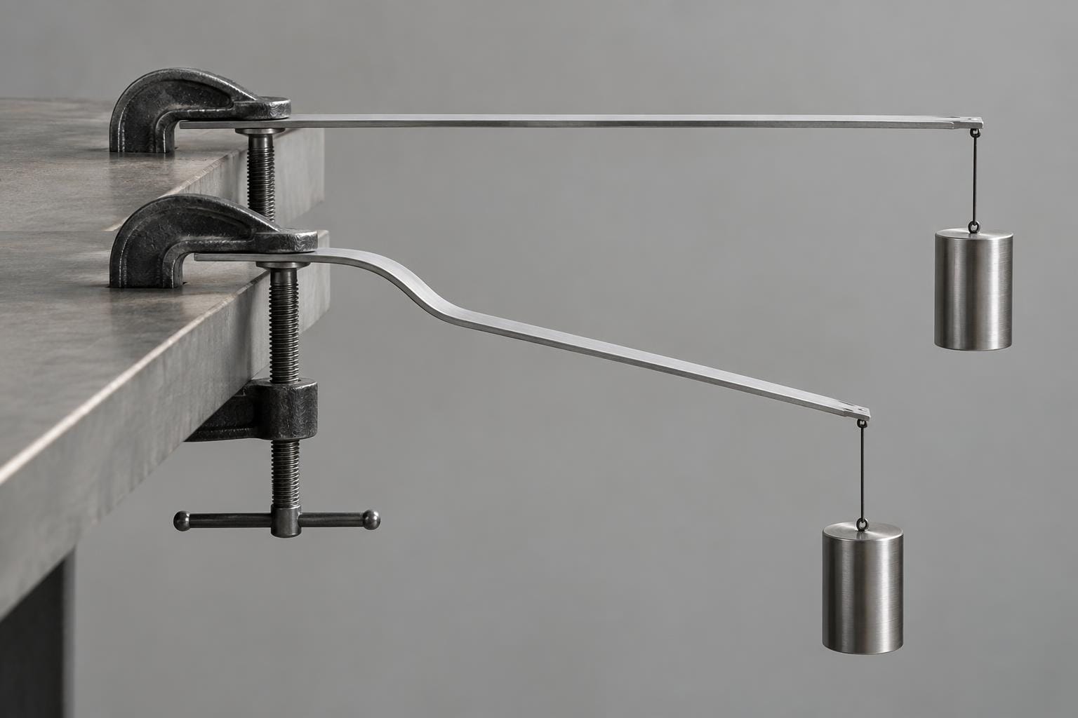 Two matching metal strips clamped to a bench carry identical suspended steel weights. One strip remains horizontal; the other buckles near its clamp and bends sharply downward.