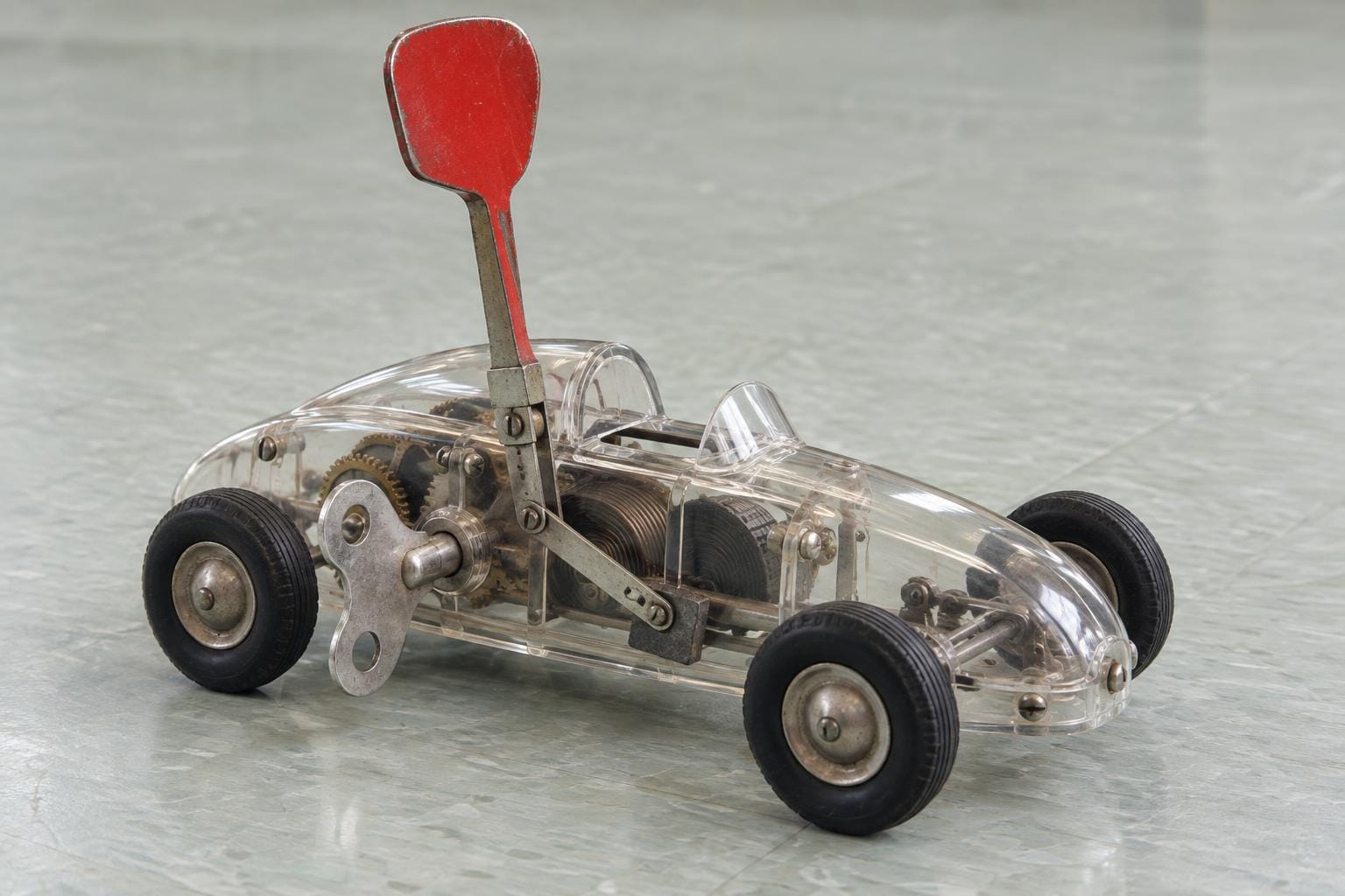 A transparent wind-up toy racing car with exposed gears and a side winding key. An oversized red stop lever towers above its roof, connected by a visible metal linkage to a brake at the rear…