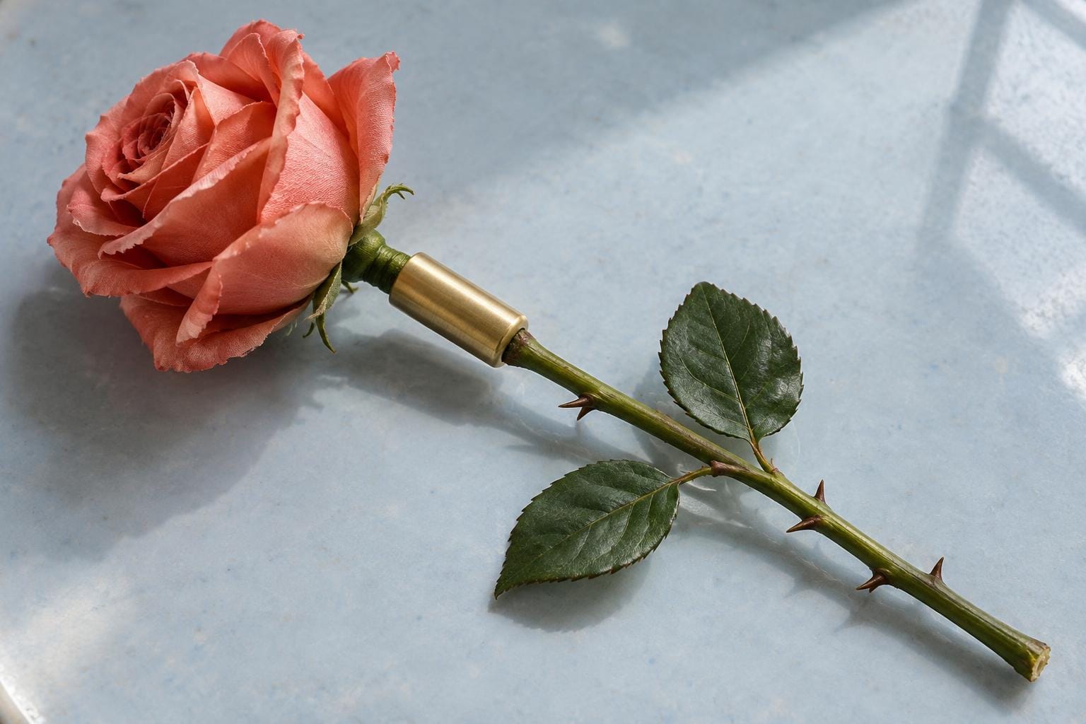 A coral silk rose lies diagonally on a pale blue surface. An exposed brass sleeve joins its short artificial stalk to a fresh green stem with leaves and thorns.