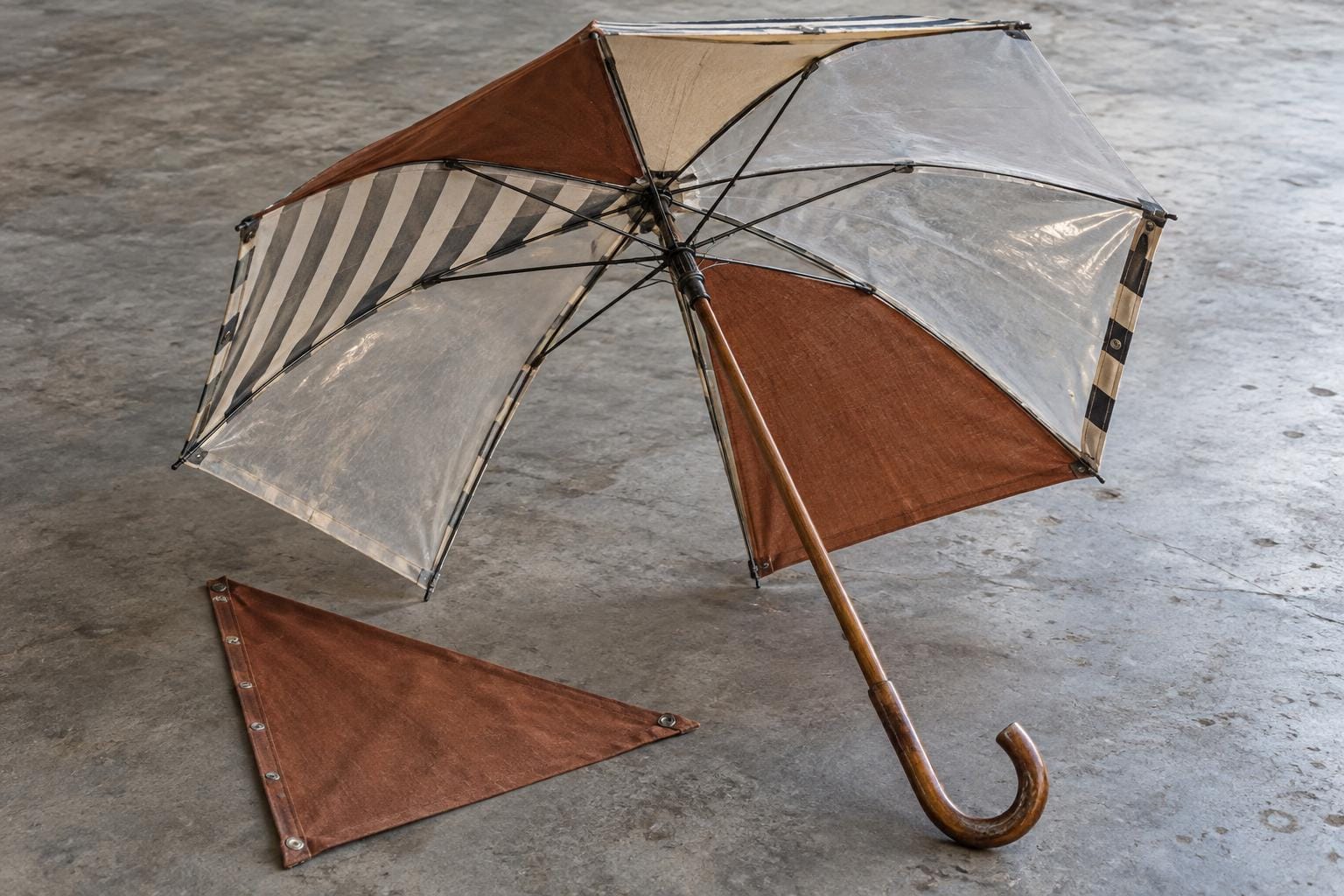 An open umbrella with striped, rust-colored and translucent panels rests on its rim. One broad triangular panel is missing, exposing the intact ribs and central hub; the detached cloth lies…
