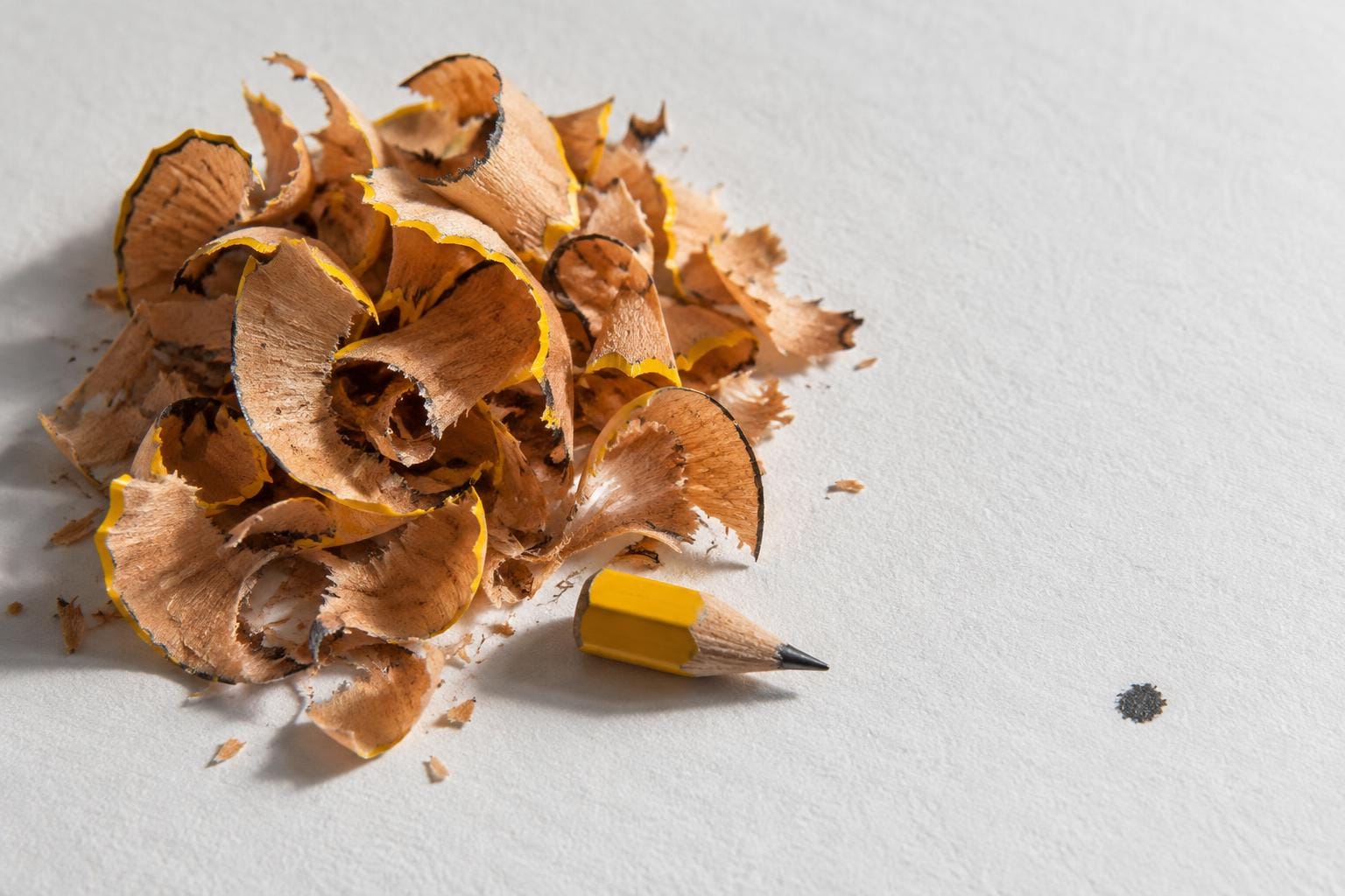 A tiny yellow pencil stub rests beside a mound of curled wood shavings on white paper. A single dark graphite dot sits apart on the right.