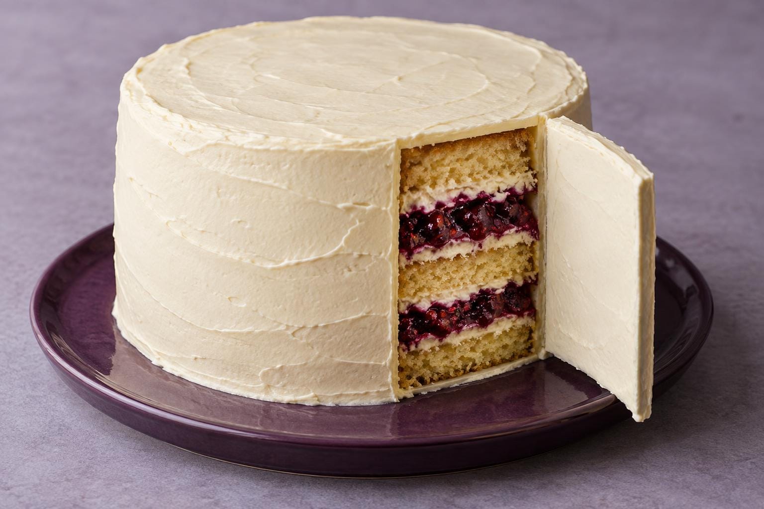 A whole ivory-iced cake has a curved shutter swung open in its side, exposing golden sponge, pale cream and dark berry layers beneath its intact top.