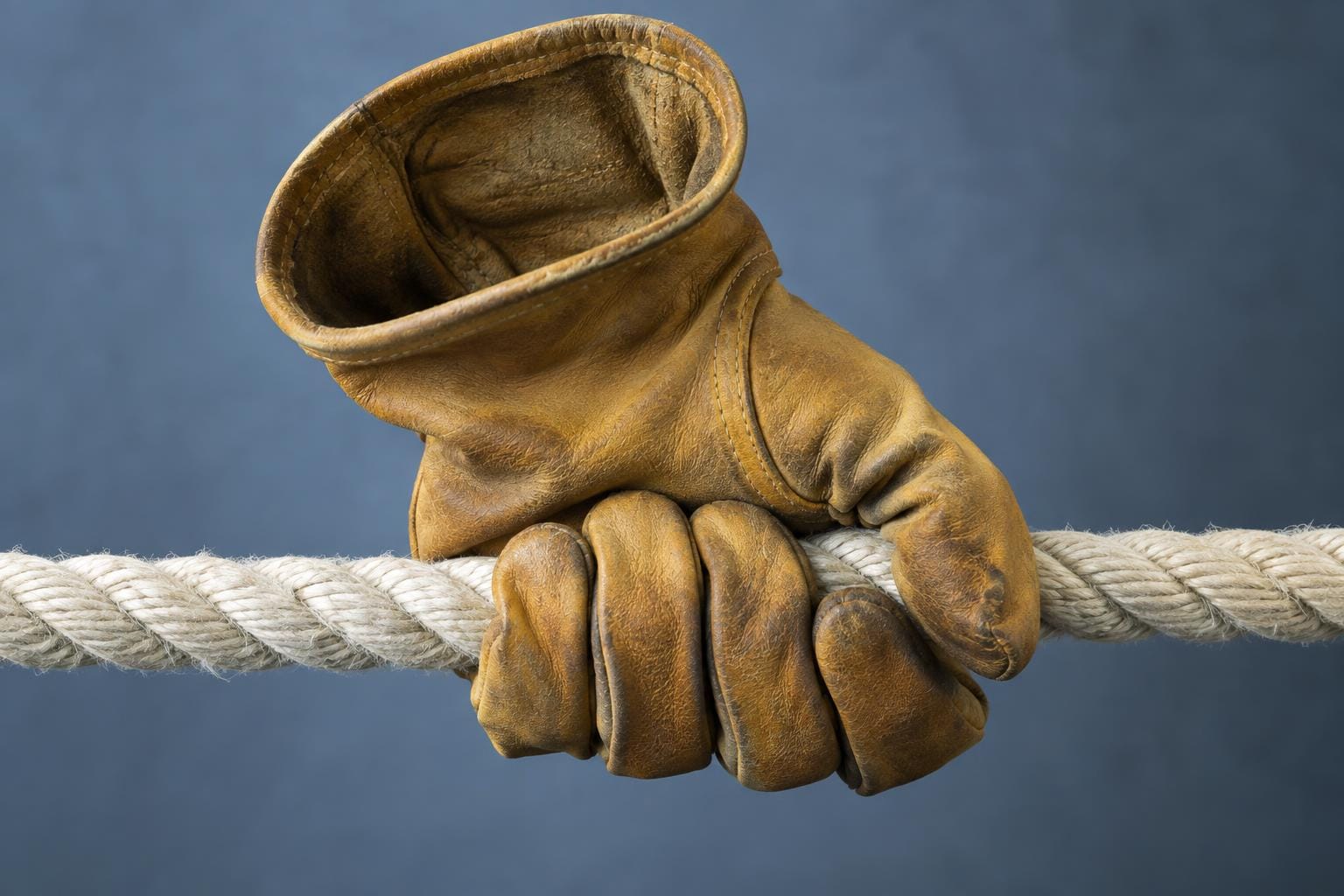 An empty ochre leather work glove grips a taut white rope. Its open cuff reveals a hollow interior while its curled fingers visibly compress the rope.
