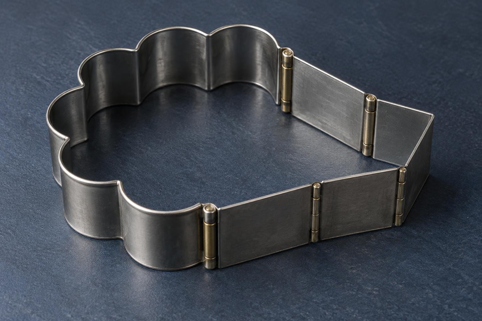 A stainless-steel cookie cutter has rounded scallops along one half and brass-hinged steel plates along the other, forming a large angular extension in its closed outline.