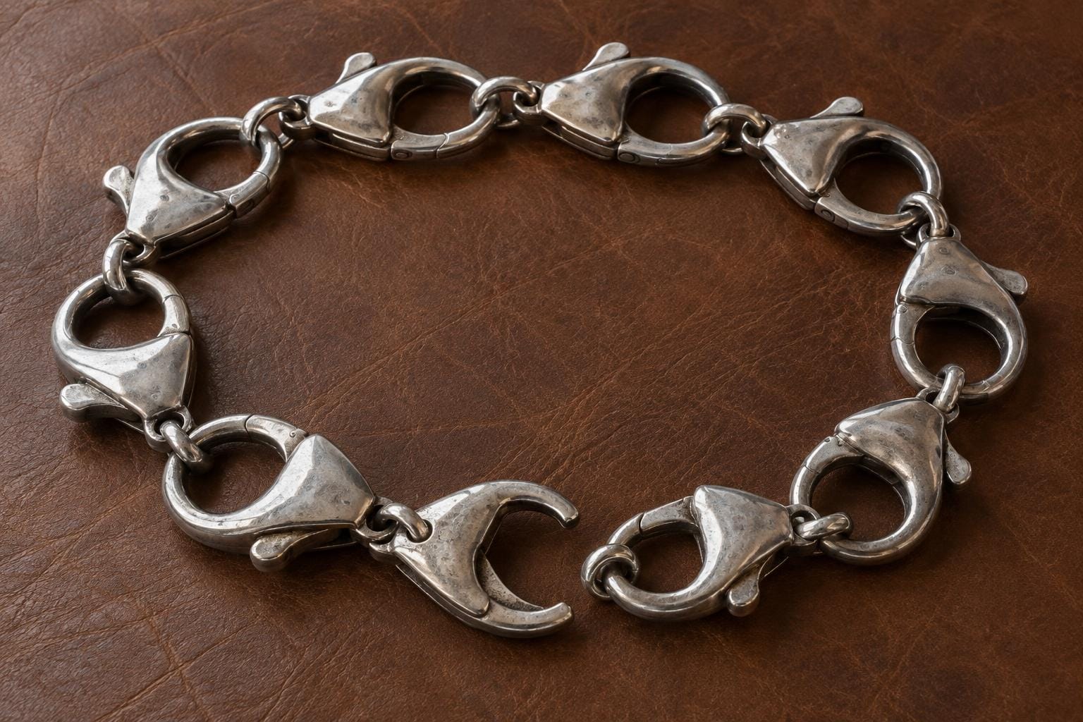 A silver necklace made entirely of large lobster clasps lies in an oval on brown leather. At the front, one clasp is open, with the neighboring clasp disconnected just outside its mouth.
